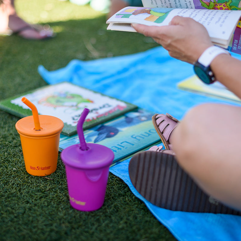 Person reading a book with colorful sippy cups on a grassy surface
