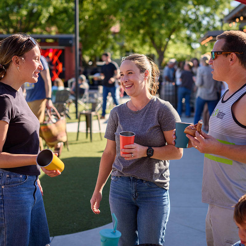 Three people standing outdoors, holding drinks, with a casual setting in the background.